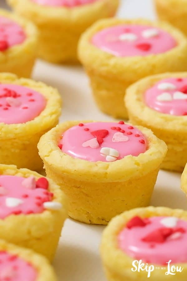 Golden yellow sugar cookies shaped like little cups, filled with pink icing and topped with red, white and pink heart sprinkles, by Skip to my Lou.