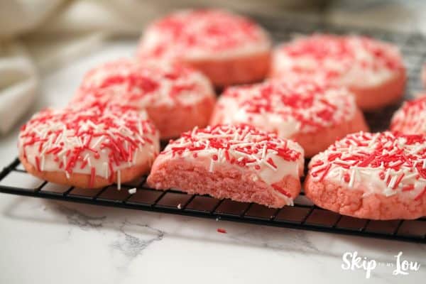 8 pink strawberry cookies with icing and sprinkles sitting on a black wire cooling rack. Half of one cookie is missing, by Skip to my Lou.