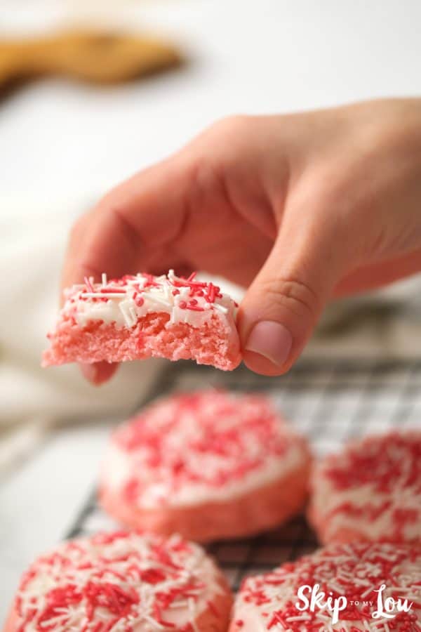 Hand holding a half-eaten pink strawberry cookie with icing and sprinkles, by Skip to my Lou.