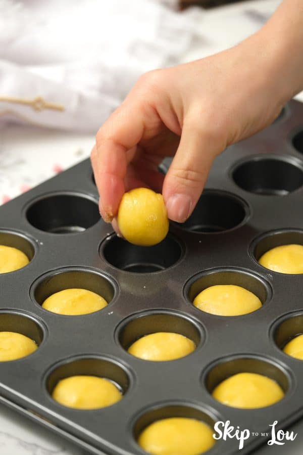 A hand placing a round ball of cookie dough into a cup of a mini muffin baking tin, by Skip to my Lou.