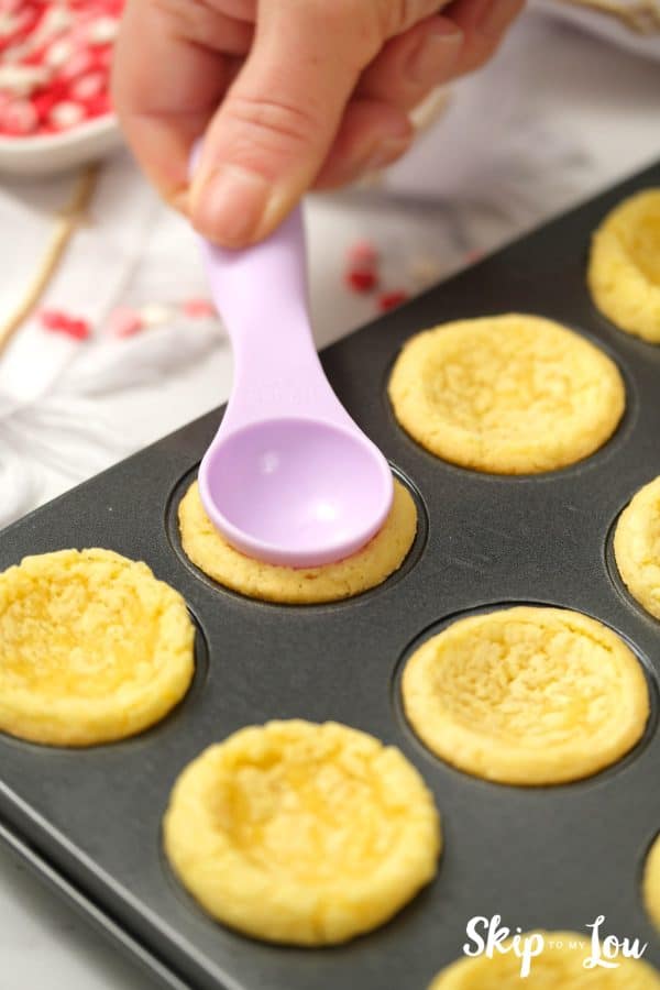 Lilac colored plastic spoon pressing down on the middle of a sugar cookie baked in a mini muffin tin, by Skip to my Lou.