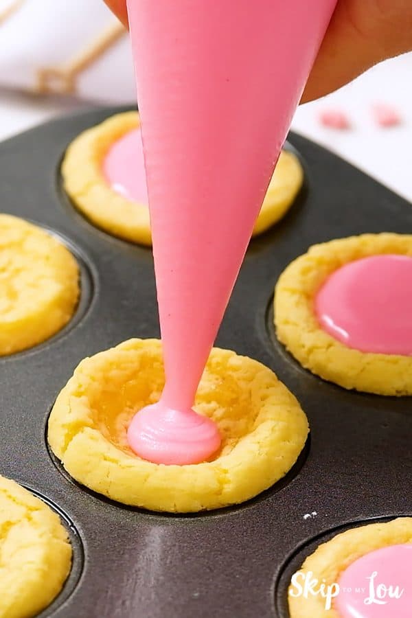 A piping bag filled with pink icing filling the center of a sugar cookie, by Skip to my Lou.