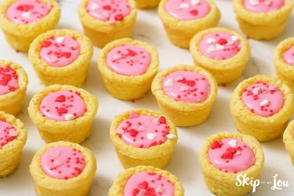 A white table with 17 Valentine sugar cookie cups sitting in rows, by Skip to my Lou.