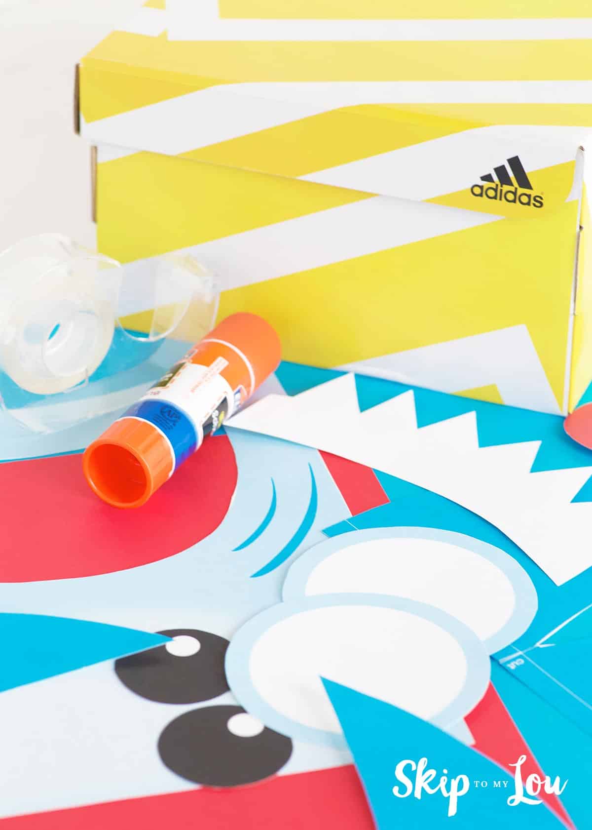 Supplies needed for making a shark Valentine box sitting out. Supplies are a yellow and white striped shoe box, clear tape roll, glue stick and colored paper by Skip to my Lou.