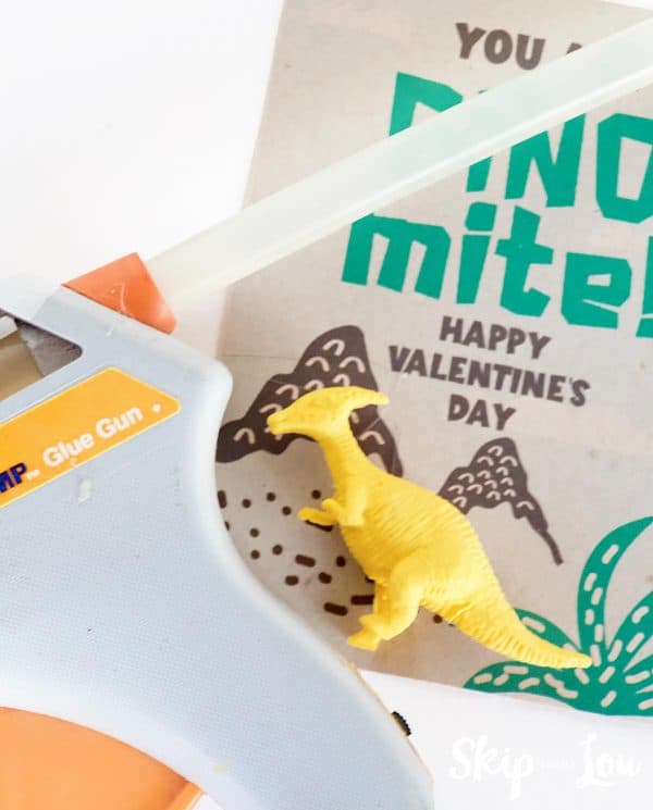 Gray glue gun with glue stick loaded. Brown card with dinosaur Valentine in the background. Yellow dinosaur hot glued on the card by Skip to my Lou.