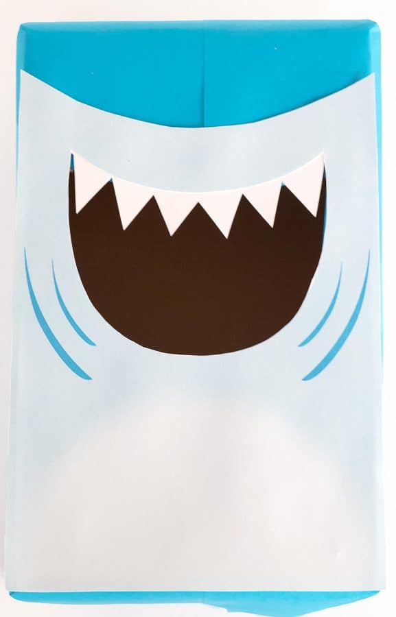 Blue box with white paper forming the shark's face and belly. Sharp white teeth hang from the shark's top lip by Skip to my Lou.