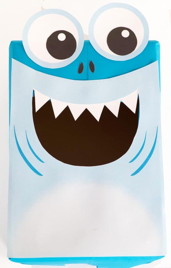 Blue box with white paper forming the shark's face and belly. Large black and white eyes, 2 black nostrils and sharp white teeth hang from the shark's top lip by Skip to my Lou.