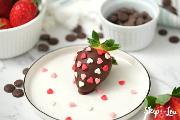 One chocolate dipped strawberry covered in red, white and pink candy hearts, sitting on a white plate, by Skip to my Lou.