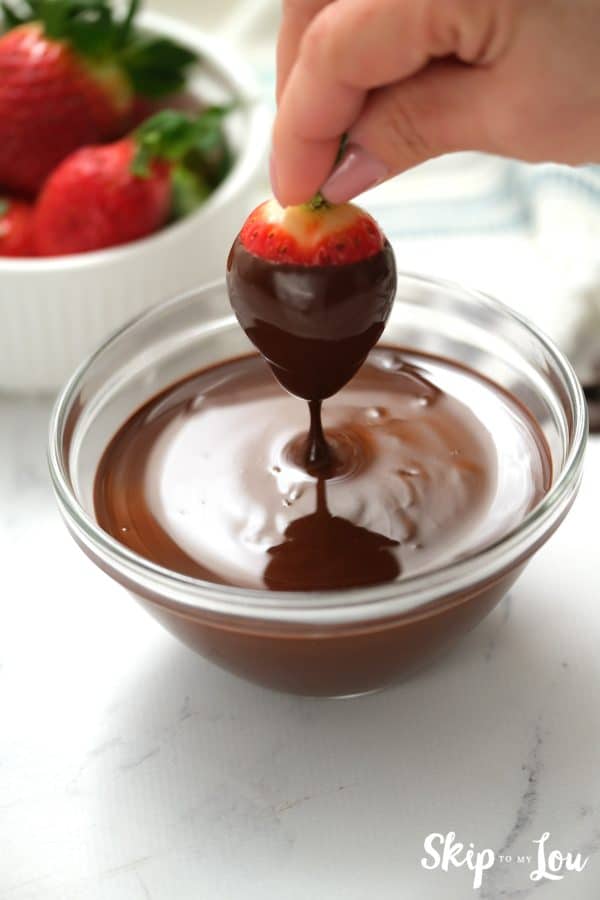 Fingers holding chocolate covered strawberry dripping into a bowl of melted chocolate, by Skip to my Lou.