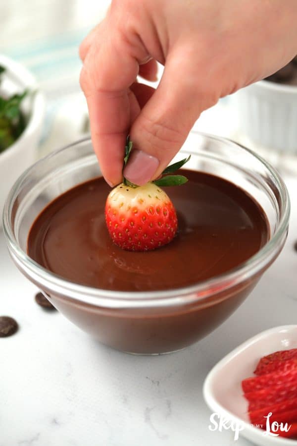 Fingers holding the leaves of a strawberry being dipped into melted chocolate.