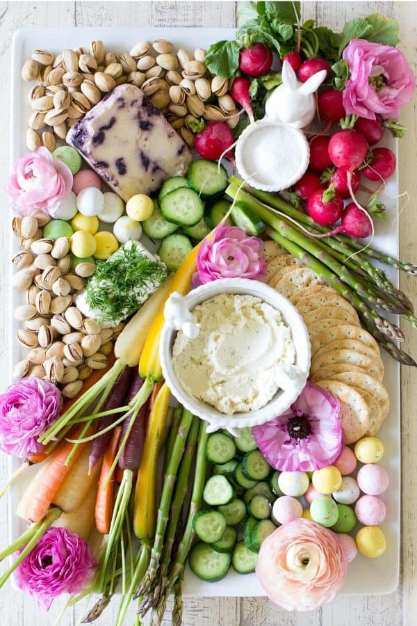 A pastel colored Easter charcuterie board idea from Freutcake that includes veggies, dips, cracker, and some flowers for decoration.
