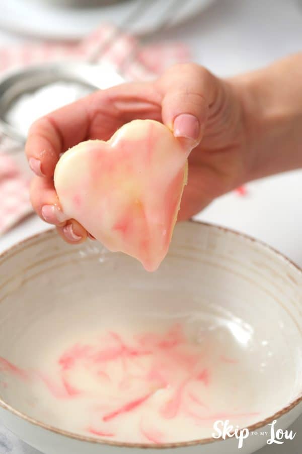 Hand holding a freshly dipped sugar cookie over a white ceramic bowl with red, pink and white confectioner's sugar icing inside, by Skip to my Lou.