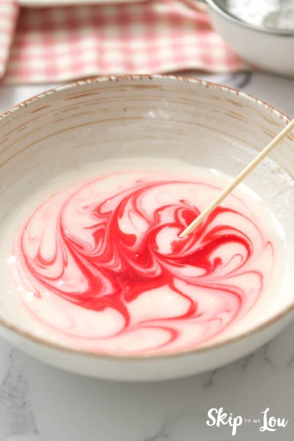 White ceramic mixing bowl with confectioner's icing and red icing gel swirled around with a wooden mixing stick, by Skip to my Lou.