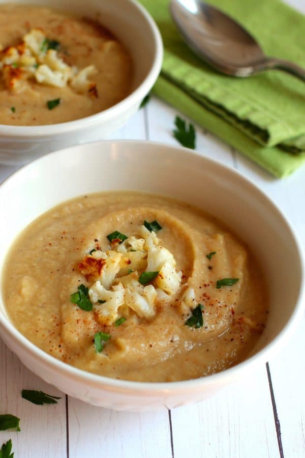 A bowl of roasted cauliflower soup, garnished with nutmeg, roasted cauliflower and parsley, served in a white soup bowl, by Skip to my Lou.