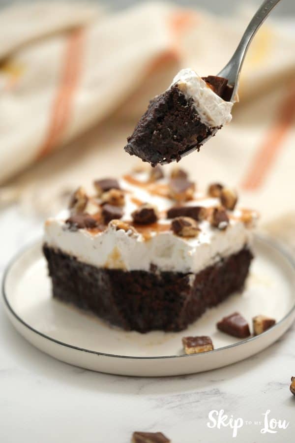 Fork holding a bite of chocolate cake with caramel sauce, topped with whipped topping and chocolate covered toffee bits, by Skip to my Lou.