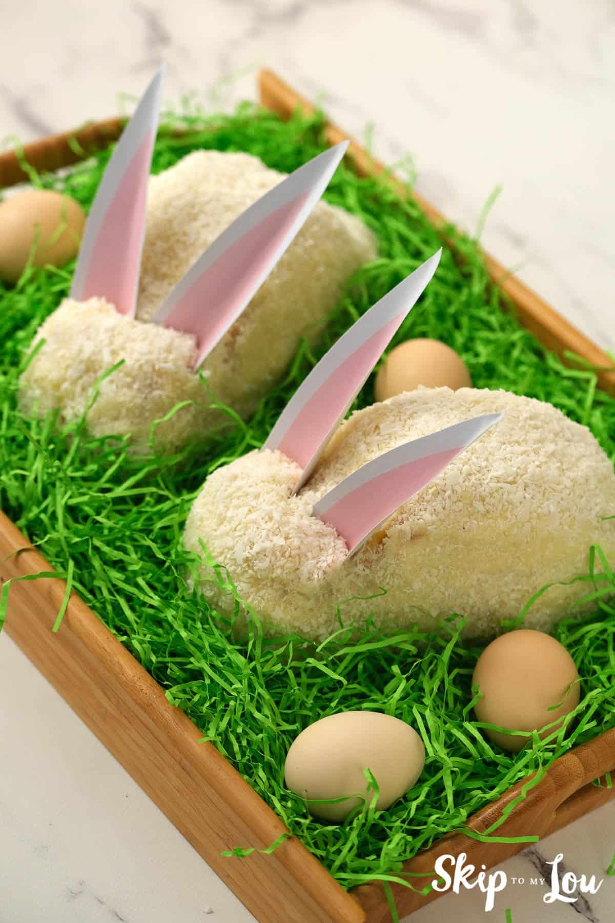 Two white Easter bunny cakes sitting on a bed of green Easter grass, by Skip to my Lou.