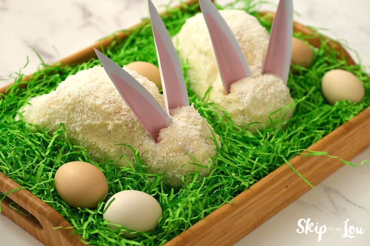 Two white Easter bunny cakes with pink and white paper ears, sitting in green Easter grass with 4 eggs, by Skip to my Lou.