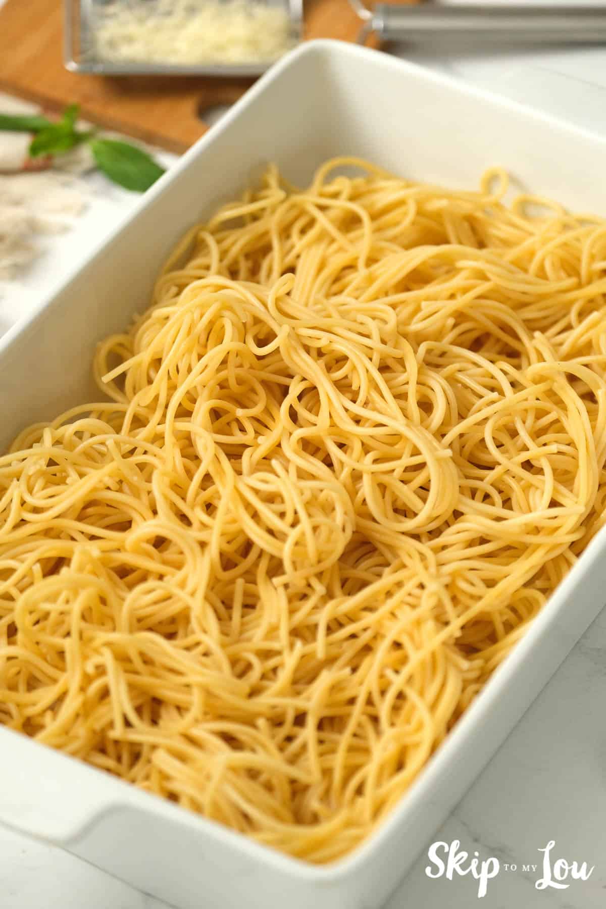 Cooked and drained spaghetti noodles in a rectangular white casserole dish, by Skip to my Lou.