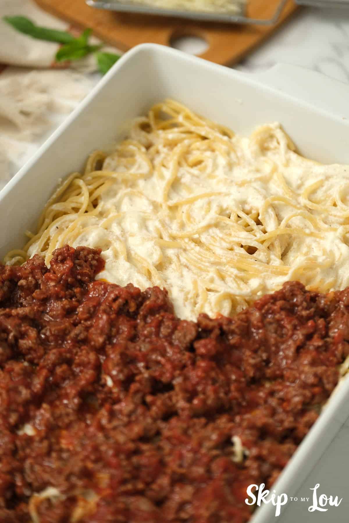 Meat sauce covering Alfredo spaghetti in a white casserole dish, by Skip to my Lou.