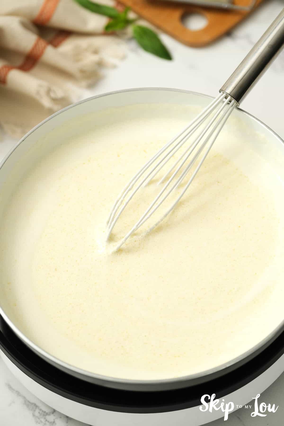 Homemade Alfredo sauce with butter, heavy cream and seasonings being whisked in a pan, by Skip to my Lou.