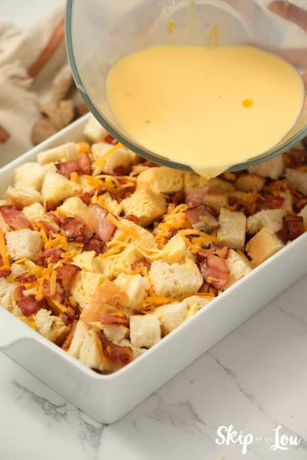 egg mixture pouring from mixing bowl over ingredients in casserole dish for egg strata from Skip to My Lou recipe