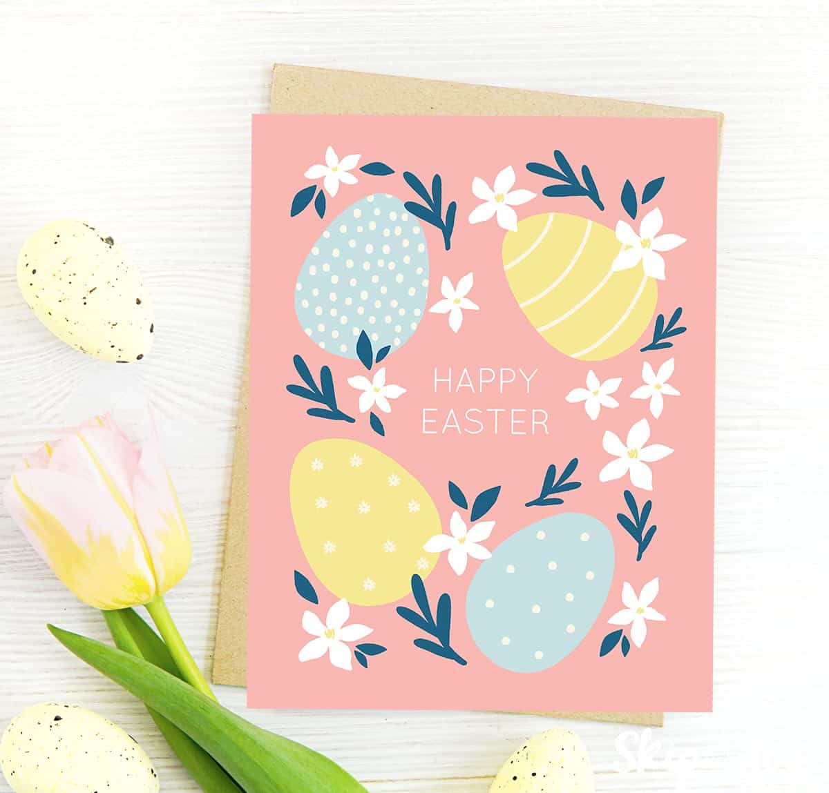Image shows a pink Happy Easter greeting card decorated with Easter eggs and flowers, next to a white flower. from skip to my lou