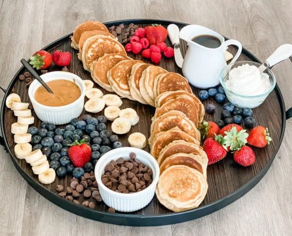 A row of pancakes surrounded by strawberries, blueberries, and bananas with whipped cream, and peanut butter from The pound dropper. 
