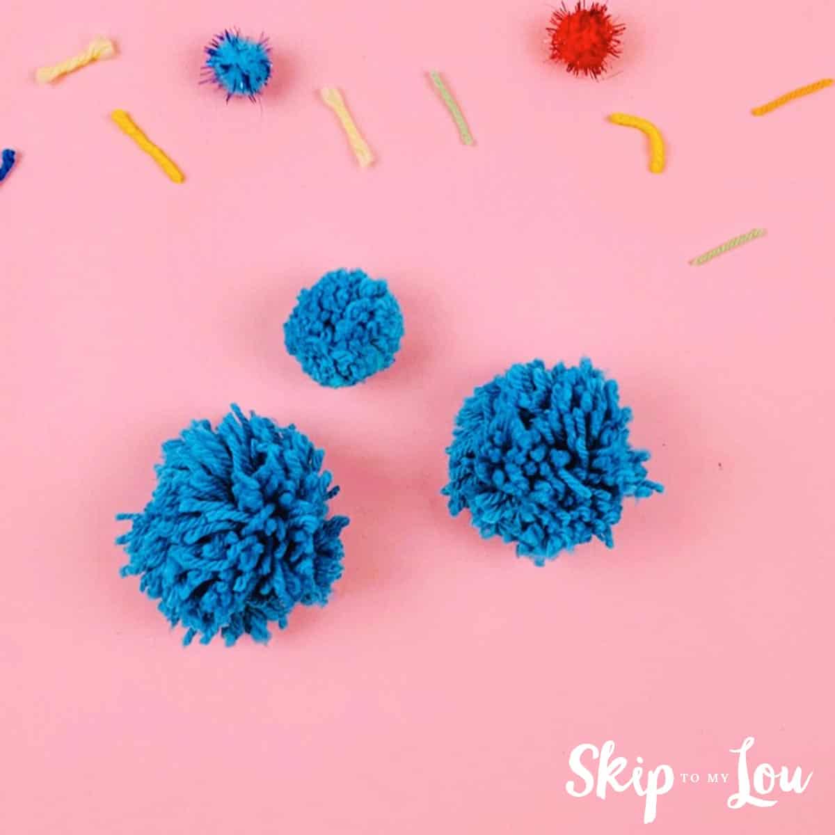 Image shows three pom poms made with blue yarn on top of a pink background, with bits of yarn around.