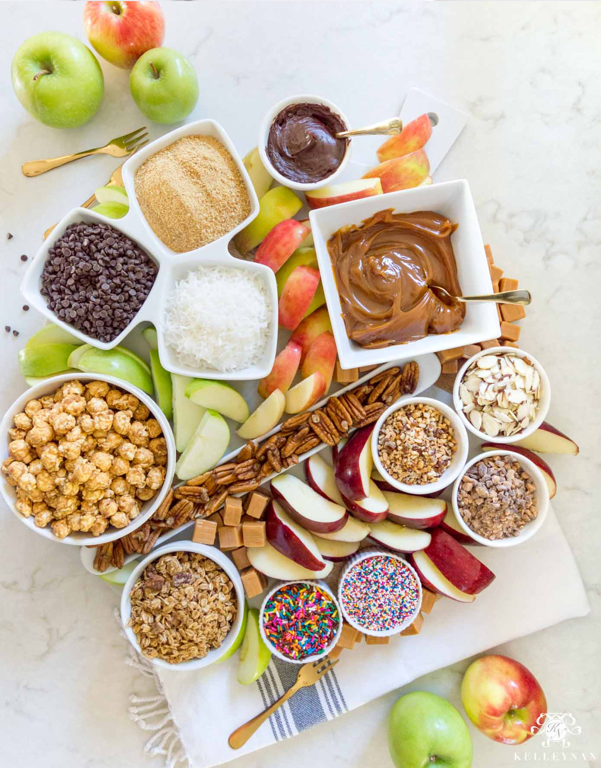Kelley Nan's Caramel Apple board presents caramel sauce, apples, chocolate chips, popcorn, crushed graham crackers, and nuts.