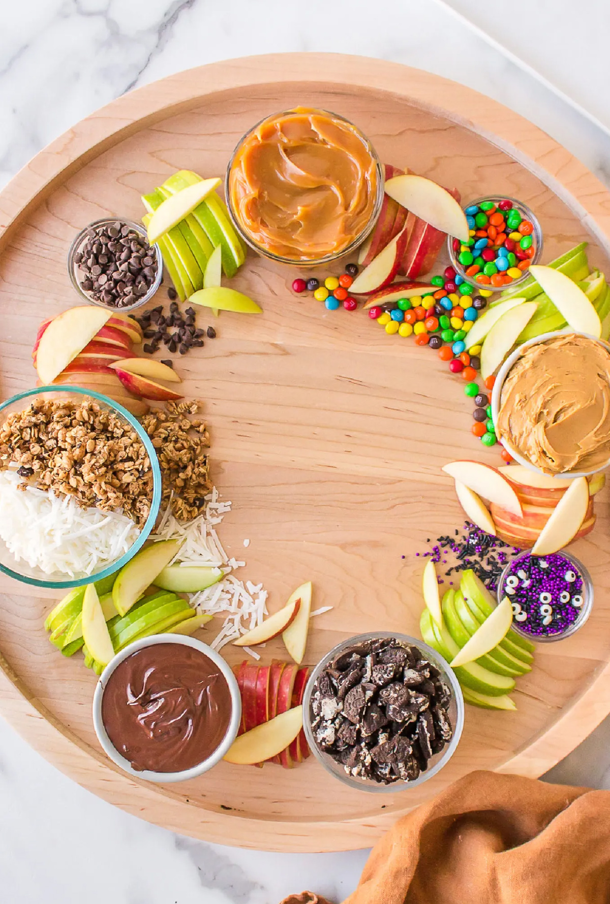 Reluctant Entertainer's Caramel Apple board showcases apple slices, m&m's, chocolate chips, caramel sauce, sprinkles, and coconut.