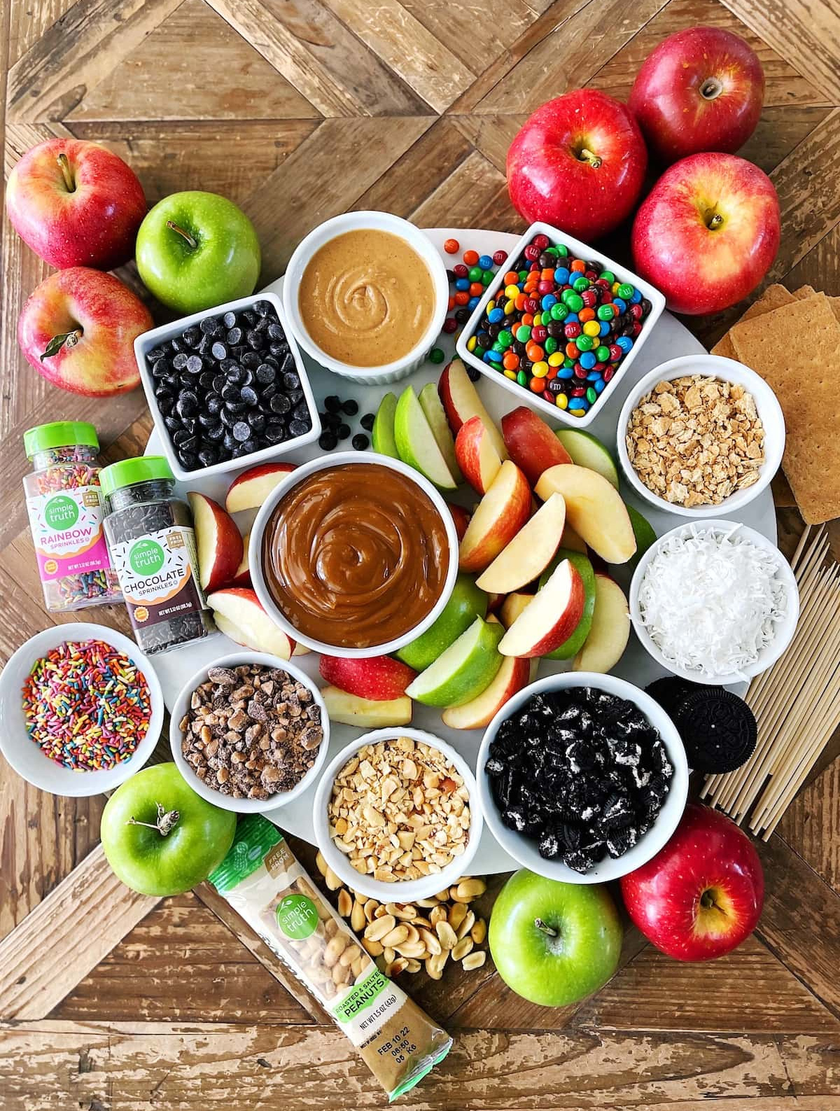 Two Peas and Their Pod's Caramel Apple board includes apples, mini m&m's, chocolate chips, sprinkles, nuts, and coconut.