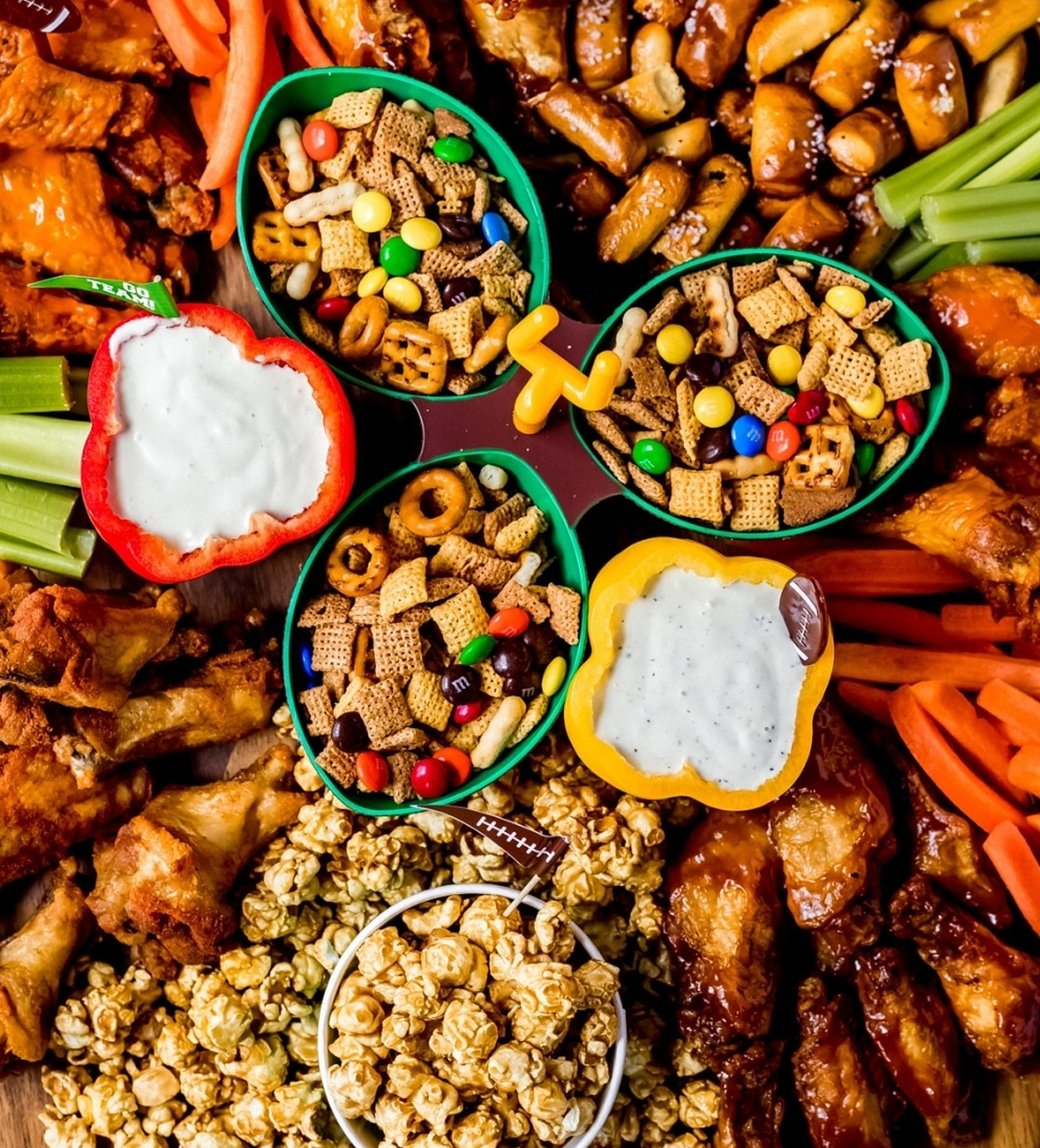 Chex mix with m&ms, popcorn, wings, dips, and veggies are displayed on Wanderlust and Wellness's football themed board.