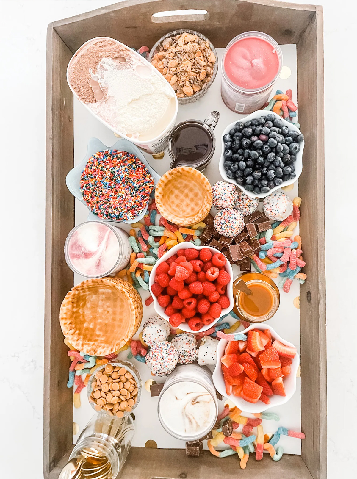 A large ice cream charcuterie board idea with plenty of broken candy bars, sour gummy worms, waffle bowls, nuts, and plenty of ice cream from Heather Krout.