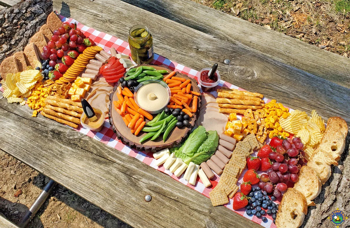 a charcuterie board laid on a picnic table with veggies, bread, meat, and cheese from let's camp more.