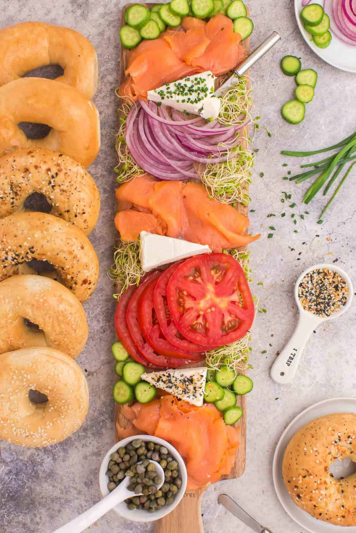 Pancake recipe's board with lox, cream cheese, cucumbers, and sprouts on a thin long cutting board with bagels surrounding.