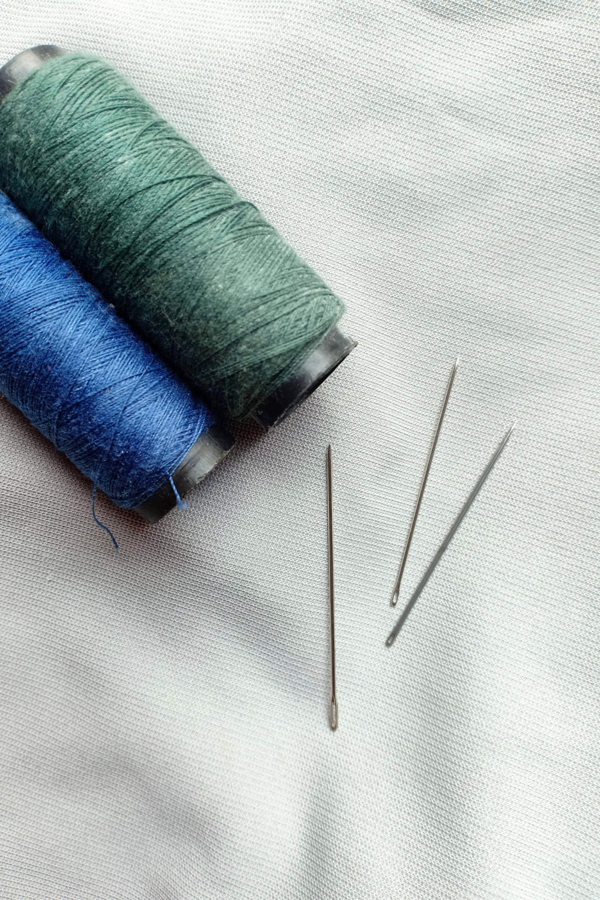 Image shows hand sewing needles next to a green thread over a grey background.
