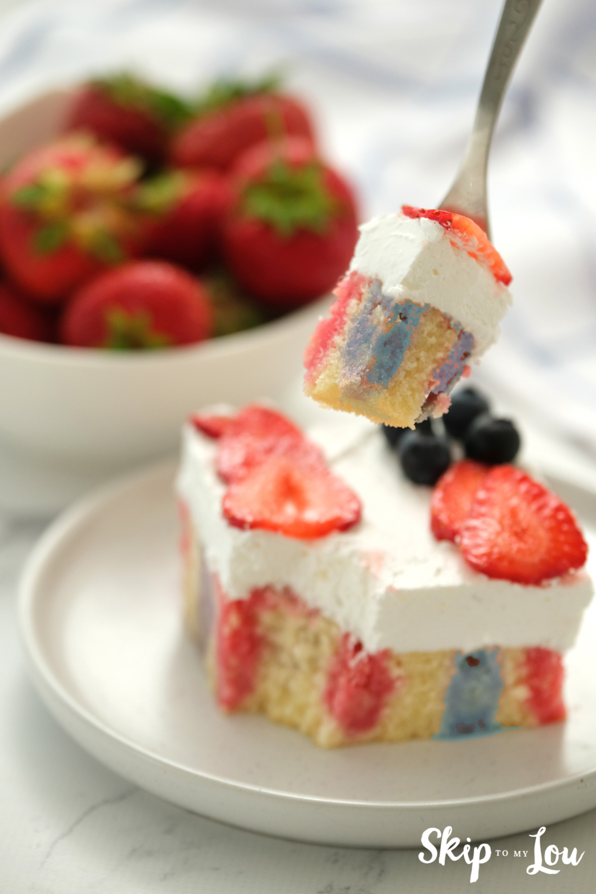 A piece of red white and blue jello poke cake with berries on top and a serving of strawberries next to it.
