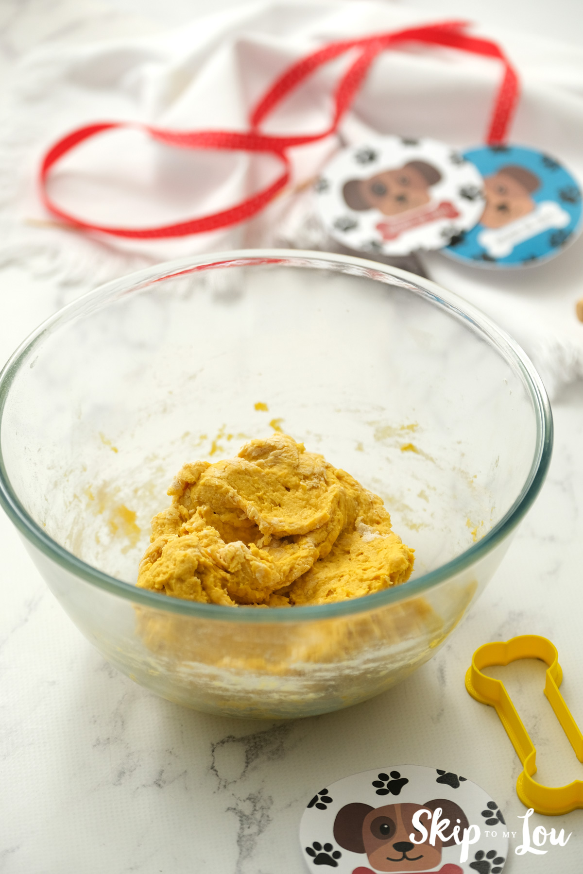 Dough for dog biscuits in a large glass bowl and a yellow dog bone shaped cookie cutter, by Skip to my Lou.