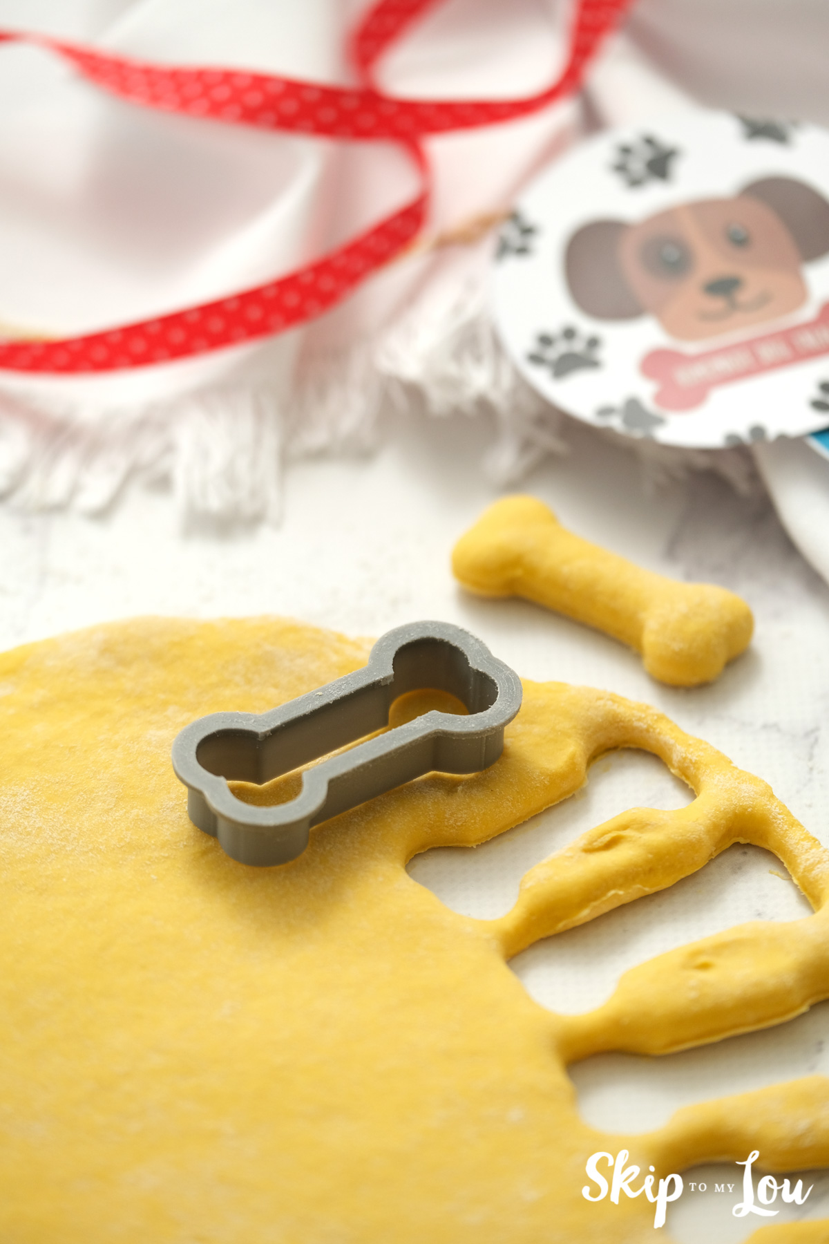 Bone shaped cookie cutter on top of dough with a dog biscuit sitting beside the dough, by Skip to my Lou.