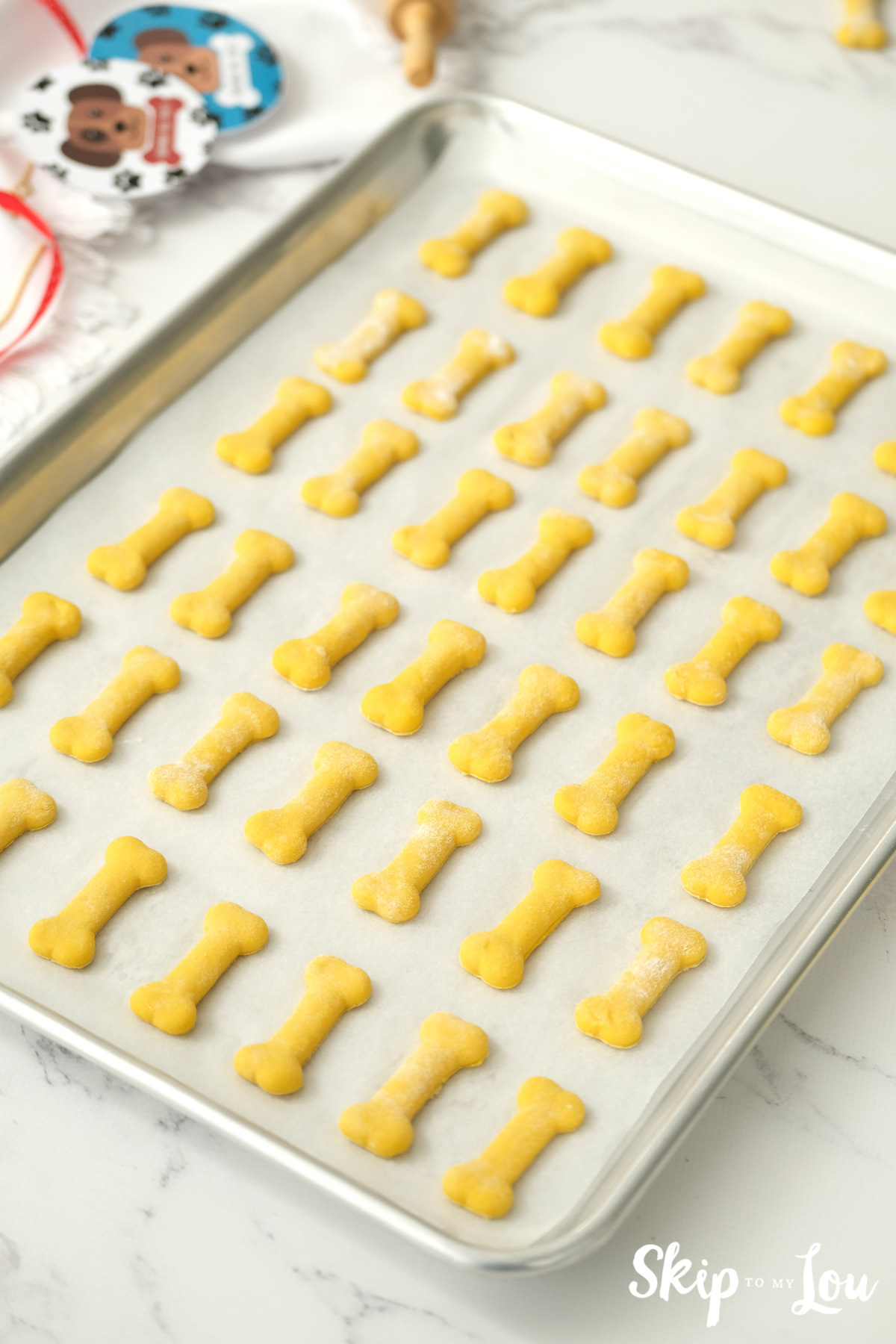 About 40 dog bone shaped dog treats on a silver baking sheet getting ready to bake, by Skip to my Lou.