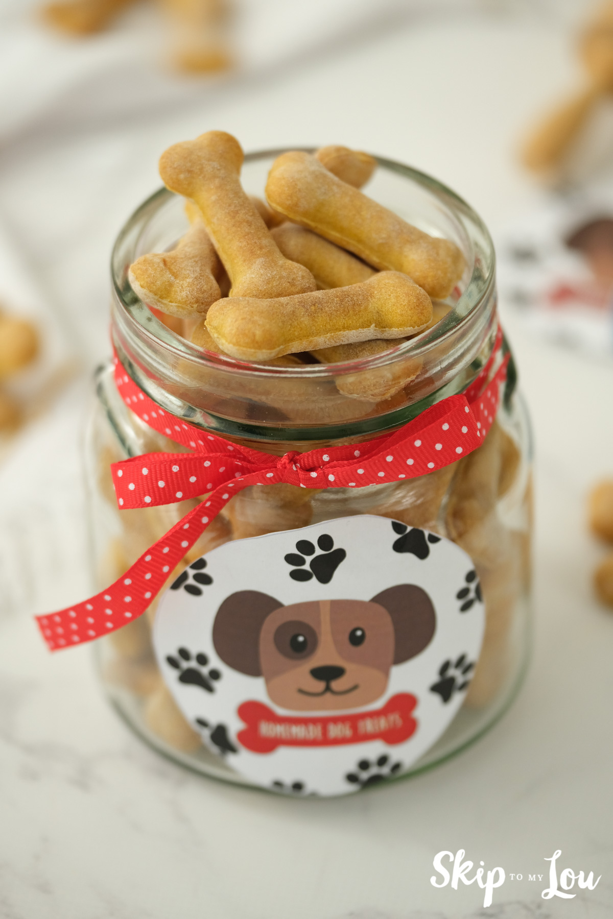 Homemade dog biscuits shaped like bones in a clear glass jar with a red polka dot ribbon and puppy label, by Skip to my Lou.