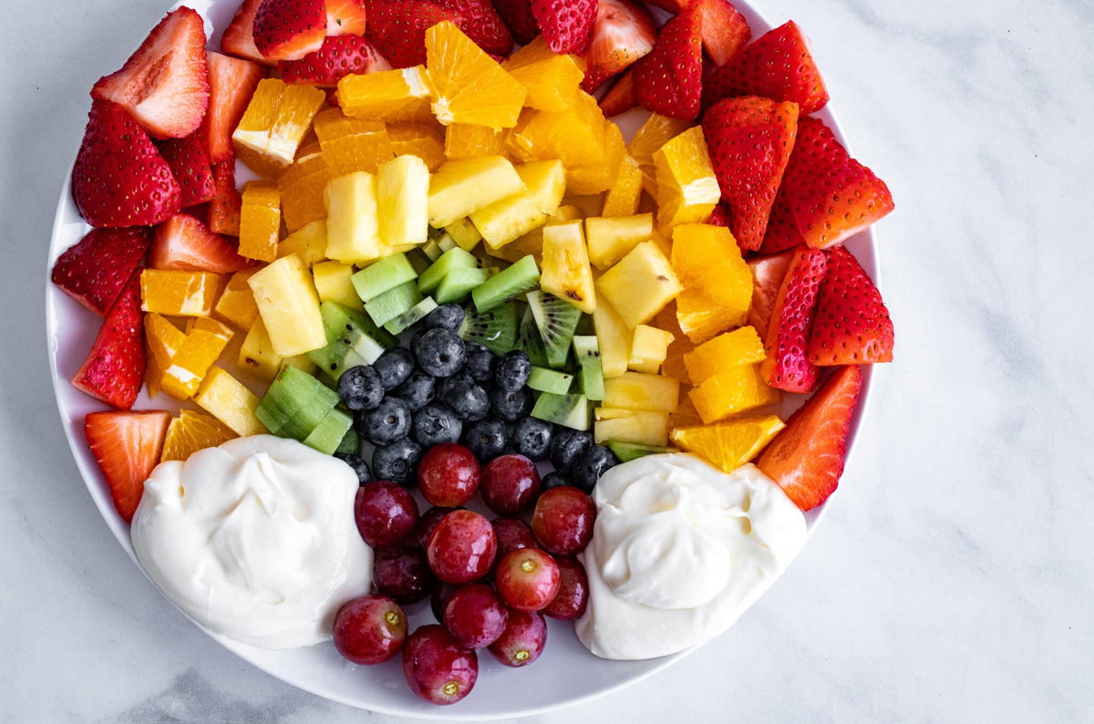a small personal rainbow board with strawberries, pineapples, grapes, and whipped cream from a red spatula.