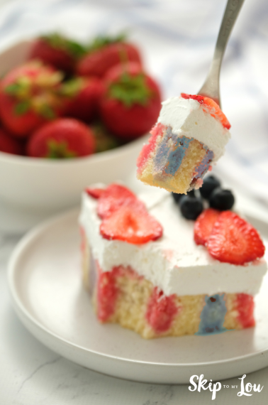 A piece of red white and blue jello poke cake with berries on top and a serving of strawberries next to it.