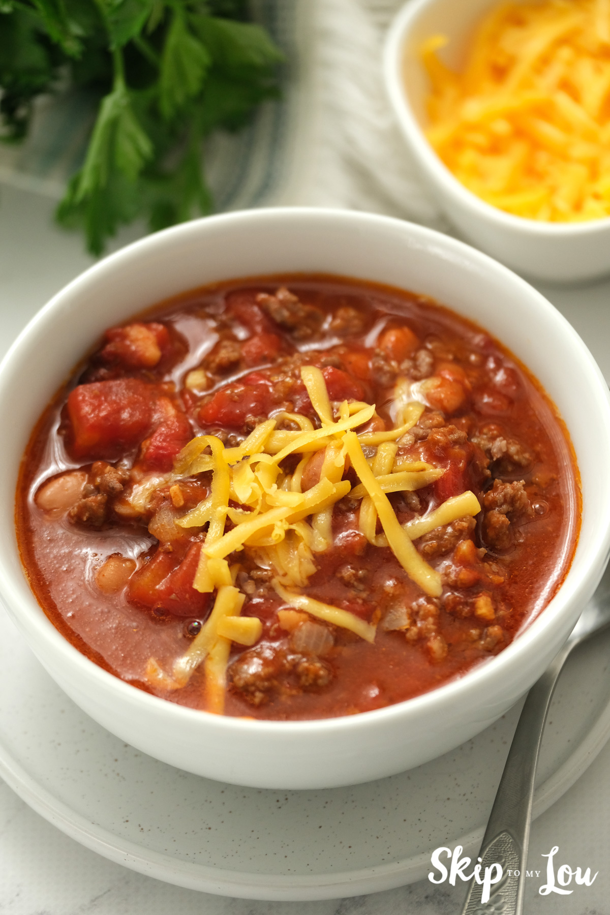 Traditional slow cooker chili in a white bowl with cheddar cheese shreds on top, by Skip to my Lou.