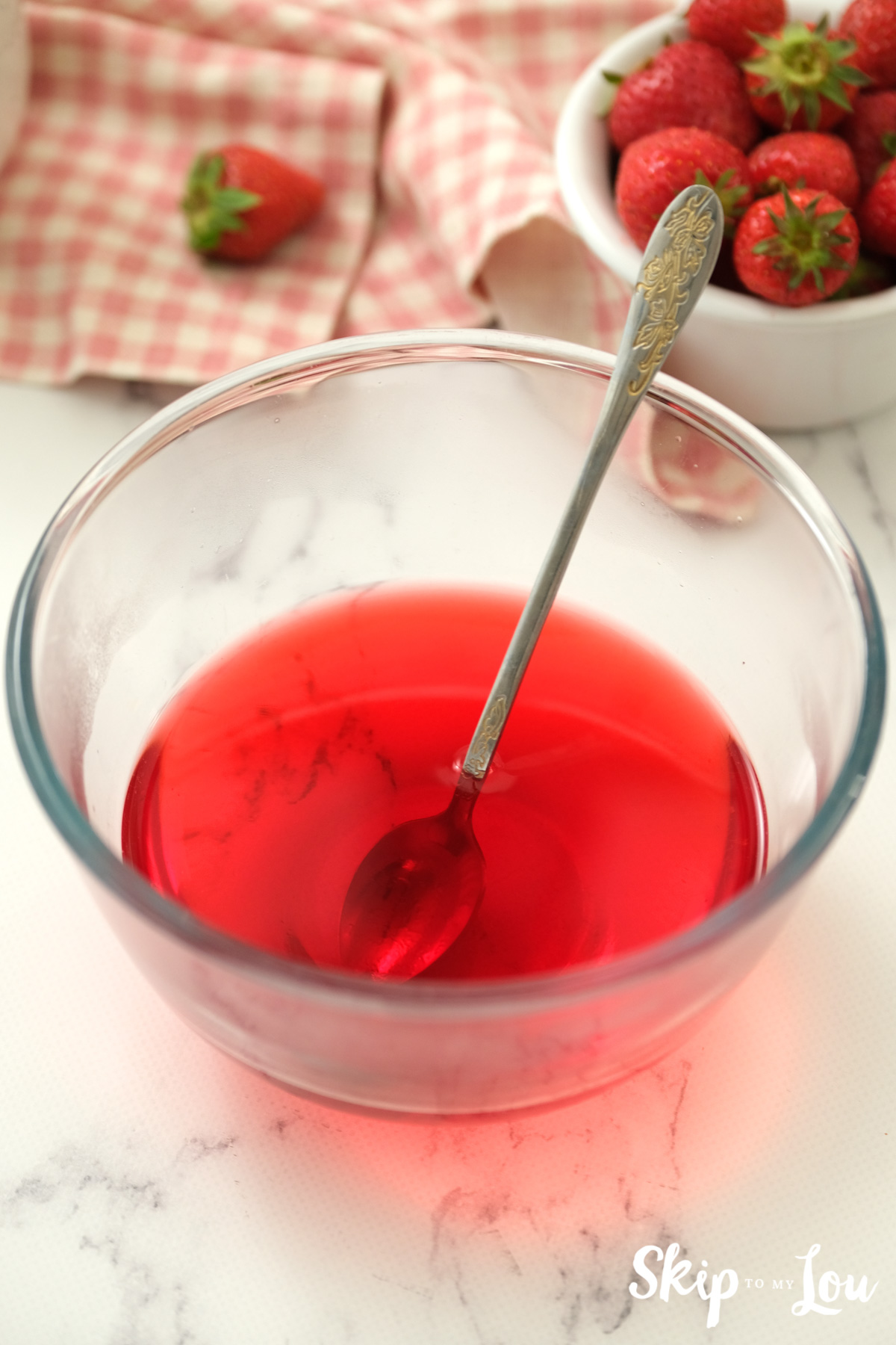 Strawberry jello and water mixed with a long silver spoon in a clear glass bowl, by Skip to my Lou.