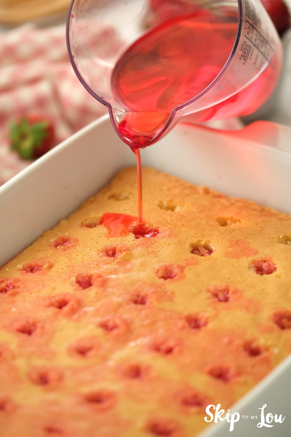 Liquid strawberry jello being poured into the holes on top of a baked cake, by Skip to my Lou.