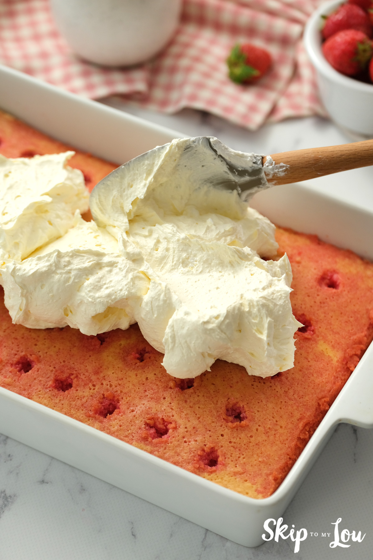 White icing spreading on top of the strawberry poke cake, by Skip to my Lou.