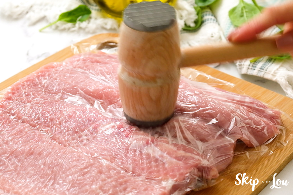 Pork loin on a wooden cutting board being pounded by a meat mallet.
