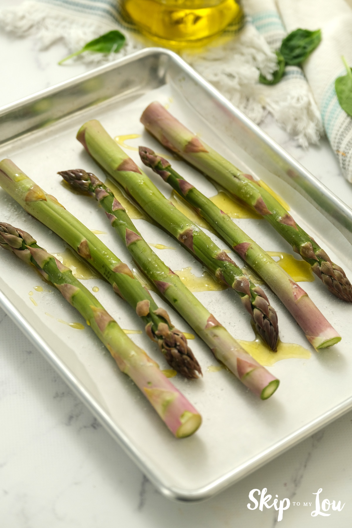 Six asparagus spears drizzled with olive oil and sprinkled with kosher salt sitting on a baking tray, by Skip to my Lou.