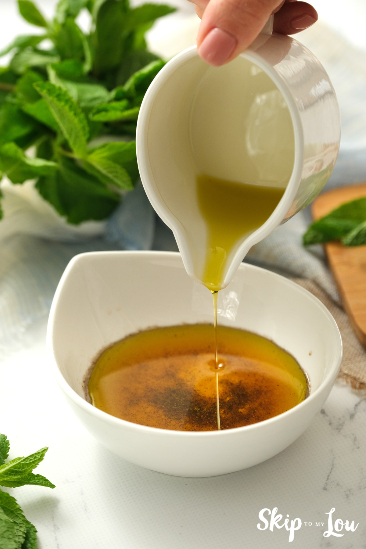 Olive oil pouring into a white bowl with herbs and spices, by Skip to my Lou.